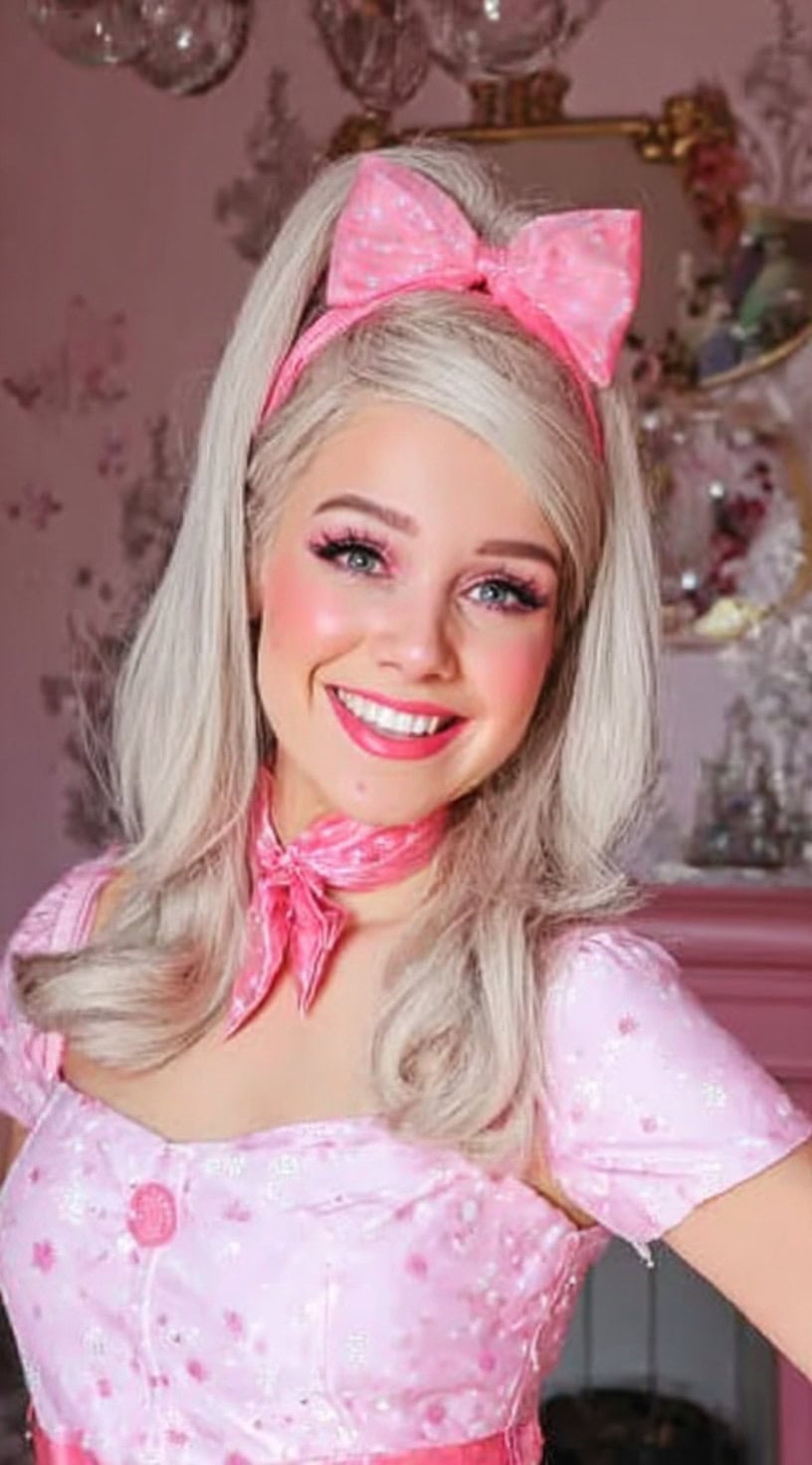 Uma mulher sorridente com cabelos loiros leves, estilizada pela metade, com pontas volumosas e invertidas, cobertas com uma faixa de cabeça de boraco de bolinhas rosa e lenço combinando.