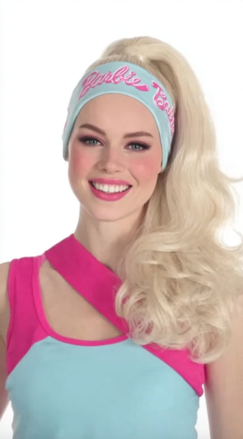 Uma mulher sorridente com cabelos loiros platinados estilizados em um rabo de cavalo enrolado com uma bandana retro da barbie e uma roupa rosa e azul bloqueada em cores.