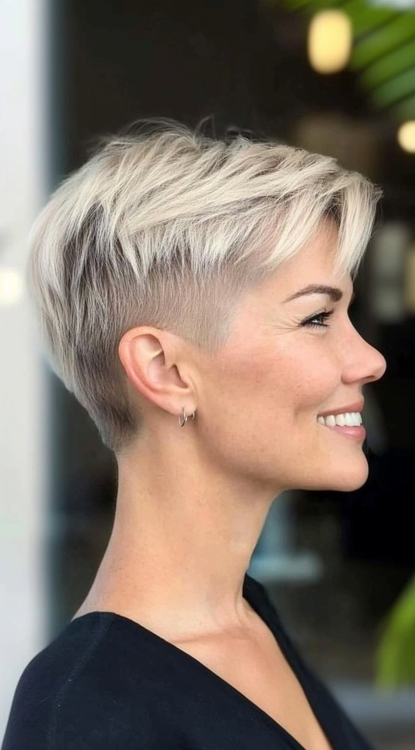 Uma mulher com um pixie loiro gelado e camadas superiores longas e varridas laterais está sorrindo de perfil.