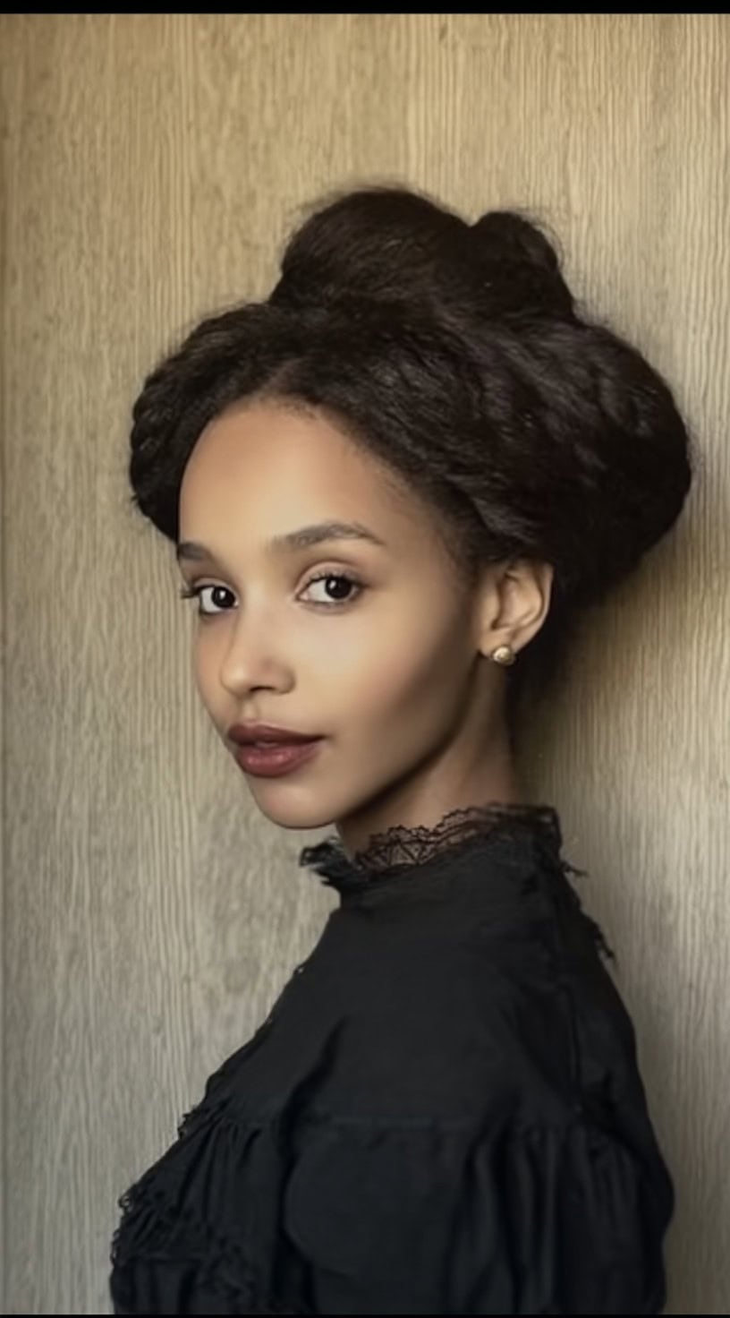 Vista de perfil de uma mulher com cabelos escuros estilizados em um grande coque torcido, combinado com um vestido preto de colarinho alto.